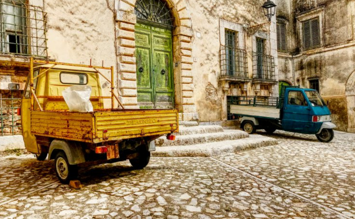 Due motocarri Ape parcheggiati in un caratteristico vicolo lastricato del centro storico, con edifici antichi e un portone verde.