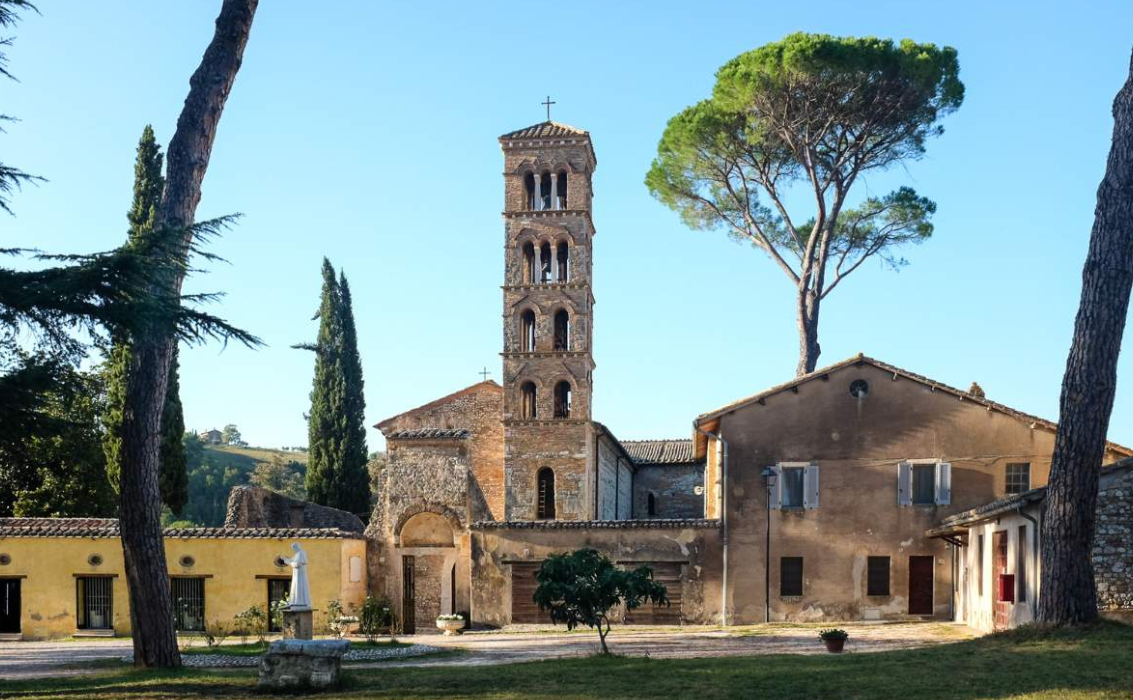 Vista esterna dell'Abbazia di Santa Maria di Vescovio a Torri in Sabina, con il suo campanile romanico e alberi circostanti sotto un cielo azzurro.